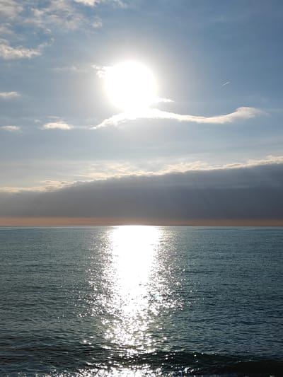 The media file displays a serene seascape, likely captured from the coast of Kobuleti, Georgia. The scene is dominated by a brilliant sun high in the sky, positioned slightly to the right of the frame's vertical center, casting a powerful, sparkling reflection path across the surface of the water towards the viewer.

The sky occupies the upper two-thirds of the image, featuring a light blue hue with scattered white and grey clouds. A distinct, long, horizontal band of darker, denser clouds stretches across the middle of the sky, just above the horizon, partially obscuring the light from the sun in some areas. Higher up, to the right of the sun, a faint white contrail from an aircraft is visible against the blue sky.

The lower third of the image is filled by the calm, dark blue-green water of the sea. The sun's intense glare creates a shimmering, almost solid, column of light on the water's surface, extending from the horizon down into the foreground. Individual glints of light catch on the gentle ripples and small waves, creating a mosaic of brightness. A subtle, reddish-orange band of light is visible along the distant horizon, separating the dark clouds from the sea, possibly indicating distant land or an atmospheric effect during sunrise or sunset.

No people or artificial objects other than the distant contrail are discernible. The overall impression is one of natural beauty and tranquility, emphasizing the vastness of the sea and sky under a bright, dynamic sun. There is no visible text in the image.