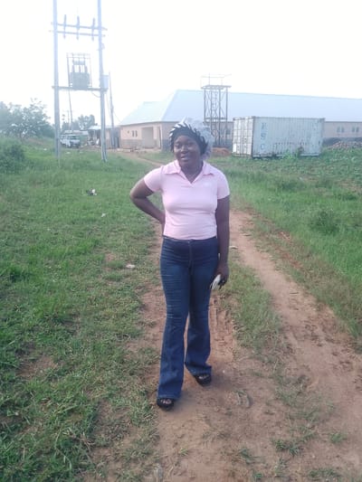 A young woman stands on a dirt path in Agwala, Nigeria, with a building and a utility pole in the background. She is wearing a light pink polo shirt with a logo and dark blue flared jeans, along with a matching headscarf. Her left hand is on her hip, and she is looking at the camera with a slight smile. A phone or small object is visible in her right hand. The setting appears to be outdoors, with green grass and a dirt path. In the background, a building with a white roof and a large white shipping container with the letters "FSC" are visible. A tall utility pole with electrical equipment is also present. The overall lighting suggests it is daytime, possibly late afternoon. The scene is calm and casual.