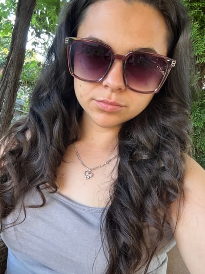 A woman is captured in a close-up selfie outdoors. She has long, dark, wavy hair and is wearing oversized, square-shaped sunglasses with a pinkish tint. A silver chain with a heart-shaped pendant rests on her grey tank top. The background consists of lush greenery, including trees and bushes, suggesting a natural, possibly garden setting. The lighting indicates it is daytime, with sunlight filtering through the foliage. The woman appears to be relaxed, with a neutral expression. The location is identified as Oreshak, Bulgaria, a village known for its crafts and traditional way of life, which could imply the setting is a private garden or a public park within or near the village.
