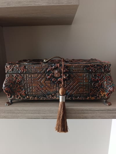 The image shows a close-up, eye-level view of an ornate, rectangular decorative box resting on a wooden shelf. The box is made of a dark material with intricate carvings and a distressed metallic finish in shades of brown, red, and teal. It features a central clasp, a raised handle on the lid, and decorative claw-like feet. A brown tassel with a silver band hangs from the front of the box. The shelf is made of light-colored wood with a visible grain. The background consists of a plain, light gray wall. There is no indication of the city of Toruń, Poland, or any urban environment in the image.