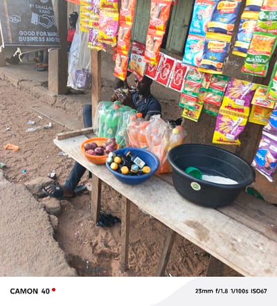 A street vendor sits behind a wooden stall displaying various goods. Packaged snacks hang above, while bottled drinks are stacked nearby. Three colorful bowls contain smaller items like onions and medicine bottles. A large black tub filled with white rice and a green scoop sits on the stall. A sign advertising "Good for Marketing, Business and..." is partially visible in the background. The surroundings show a simple, somewhat cluttered street scene.