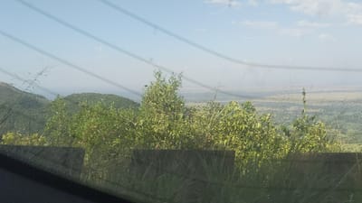Hills covered in lush green vegetation occupy the left side, transitioning to a flat expanse in the distance. The sky is pale blue with wispy clouds. A dark, blurry object is in the foreground at the bottom left. Power lines cross the upper part of the view, giving the impression of viewing from a window. Grass and a low barrier line the middle ground.