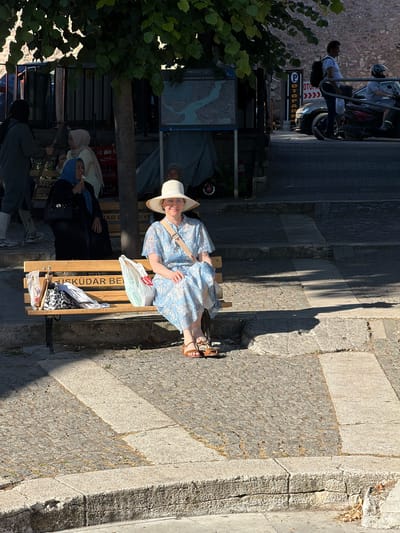 In Üsküdar, Turkey, a woman wearing a white sunhat and a light blue floral dress sits on a wooden bench. The bench has "ÜSKÜDAR BE" inscribed on it. Next to her are several shopping bags and an umbrella. To her left, another woman in a blue headscarf eats ice cream. In the background, other people are visible, along with a map displaying a waterway. The scene is set on a sunlit street with cobblestone paving.