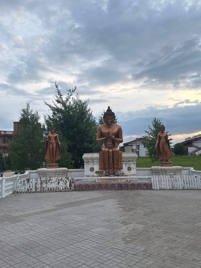 The image depicts an outdoor scene with three copper-colored statues of Buddhist figures arranged in a courtyard. The central statue is seated, while the two flanking statues are standing. The statues are placed on elevated platforms with ornate bases. In the background, there are lush green trees, buildings with white and red facades, and a cloudy sky with hints of sunset. The foreground consists of a paved courtyard made of gray interlocking bricks. A white railing with some decorative elements runs along the edge of the courtyard. The environment appears to be a public space, possibly a park or a temple complex. The city of Petrovo, Russia, is mentioned as the location, suggesting this could be a cultural landmark within the city.