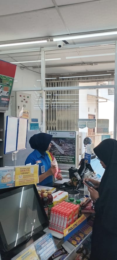 A cashier at a shop in Meunasahbaro, Indonesia, wearing a blue uniform, is checking out a customer who is wearing a black hijab and holding a phone. The checkout area is cluttered with merchandise including a stack of red boxes of "Paling Murah," Snickers candy bars, and various drinks. The cash register and a sign advertising a "Gratis" promotion are visible. Security cameras are mounted on the ceiling. A sign promoting a humanitarian effort for Sumatra is placed near the counter. Behind the cashier is the shop’s security gate.