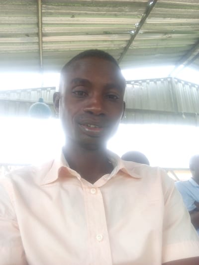 This is a medium-shot, front-facing selfie of a Black man in what appears to be an outdoor or semi-outdoor setting. He has short, dark hair and is wearing a light peach-colored collared shirt with buttons. His mouth is slightly open, and he has a gentle smile, suggesting a relaxed or pleasant emotion. To his left, a blurred, round, light-colored object hangs from above, possibly a light fixture or decorative item. Behind him, the background is bright and washed out, with a corrugated metal roof structure visible overhead. The metallic surface of the roof appears aged and somewhat weathered. There are hints of other people in the background, also out of focus. The bright light suggests it could be daytime, possibly with overcast conditions or a very bright sun due to the washed-out appearance. The overall scene has a simple, everyday feel, potentially in a community or shared living space given the "Immigration Quarters, Nigeria" location context. There is no visible text in the image.