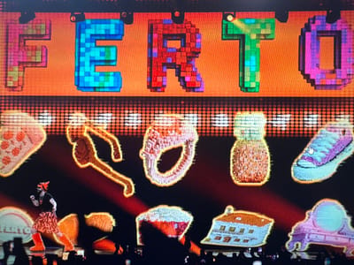 The image depicts a vibrant stage setup with large, pixelated letters spelling "FERTO" across the top. The background is a warm orange, dotted with a grid of illuminated circles. Below the letters, a series of colorful, pixelated graphics are displayed, including what appears to be a slice of pizza, a ring, a jar, and a sneaker. A performer, dressed in an orange and black tiger-striped skirt and matching fuzzy boots, with a tiger-themed headpiece, is center stage, in motion. The performer is on a stage with a dark floor, and the audience is visible at the bottom of the frame, with some people holding up phones. The scene suggests a live performance or event. Without further information, it is impossible to determine the specific location in the city of Ellwangen, Germany.
