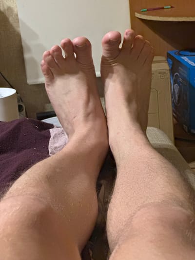 Two bare feet with visible toes and toenails are resting on a surface. The left foot is positioned slightly closer to the viewer and appears to be on a dark, textured fabric. The right foot is resting on a lighter, textured surface. The lower legs of both individuals are visible, with the left leg showing some muscle definition and the right leg covered in light hair. In the background, a white surface is visible to the left, and to the right, a cardboard box and a blue electronic device box are present. A shelf with colored pencils is also visible in the upper right corner. The scene appears to be indoors, possibly in a domestic setting, and the lighting suggests it may be evening or a dimly lit room. The location is specified as Tarasovka, Russia.
