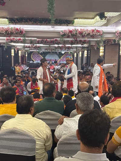 The image captures an indoor event in South Dumdum, India, within a decorated hall. The ceiling features multiple rows of pink and white floral garlands with green foliage, and various lighting fixtures. A stage area is visible in the background with a large, elaborate floral backdrop. The scene is populated by a large gathering of individuals, primarily adult males, seated in rows facing the front.
Three prominent male figures are positioned in the mid-ground, appearing to be speakers or performers. The leftmost figure wears a white kurta, a dark cap, and a red-patterned sash, facing right towards a central figure. The central figure wears a white kurta, a colorful patterned turban, and a blue-red sash, holding a microphone in his left hand while facing the leftmost figure. To their right, a third figure, wearing a white kurta and a large orange patterned sash, stands with his right arm extended outwards and slightly upwards, gesturing towards the right.
The audience members, many dressed in traditional or semi-traditional Indian attire including kurtas and head coverings, are largely seated and observing the activity at the front. A black speaker is visible on a stand on the far left. One audience member in the lower-left foreground has a mobile phone visible.