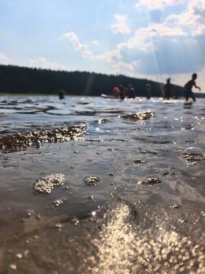 This media file depicts a group of people enjoying a sunny day at a lake, likely during the summer. The camera is positioned low to the water, offering a close-up view of the rippling surface with sunlight glinting off the waves and creating bokeh effects. In the background, silhouetted figures are visible in the water, some appearing to be children. The shoreline is lined with a dense, dark green forest, suggesting a natural, undeveloped setting. The sky is blue with scattered, fluffy white clouds, and a bright sun casts rays through the atmosphere. The overall mood is one of leisure and outdoor recreation. The time of day appears to be afternoon, given the angle of the sunlight and the bright, clear weather. The location context of Sysert, Russia, points to a potentially rural or forested area known for its lakes. There is no visible text in the image.
