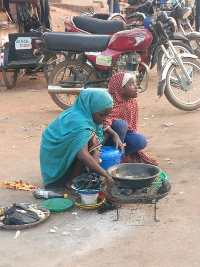 This image, captured in Alizaga, Nigeria, depicts two young females squatting on an unpaved, dusty ground, likely engaged in street vending activities. In the foreground, a female wearing a teal hijab and patterned top is actively tending to a dark, wok-like pan resting on a small, metal charcoal stove or grill. Ash is visible around the stove, indicating its use. To her left, several items are arranged on the ground: a black substance in a white bowl atop a yellow plate, a green plastic plate with liquid, an empty aluminum plate with white remnants, a dark bottle, and a folded piece of patterned fabric. A blue plastic bucket with a lid is positioned between the two individuals. The second female, to the right and slightly behind, wears a red and white patterned hijab and blue pants, looking upwards and to the right, observing the surroundings. In the mid-ground, multiple motorcycles are parked, including a prominent red motorcycle with a black seat and a sidecar-like attachment displaying "WOFAN ICON". Other blurred figures are visible in the background. The overall environment is an outdoor, earthy setting typical of a marketplace or street.