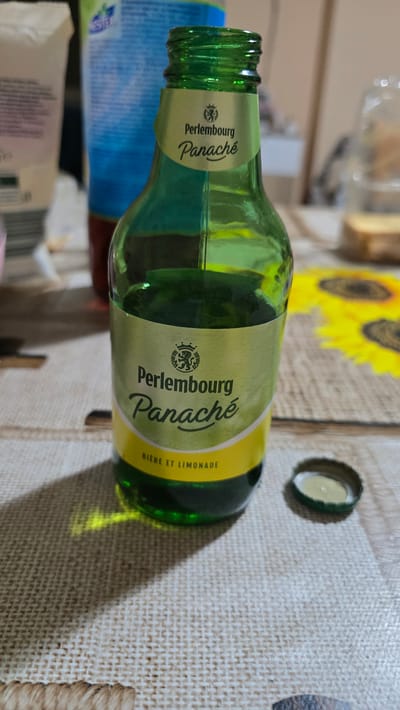 A green glass bottle of Perlembourg Panaché, a beer and lemonade beverage, stands on a table. The bottle has a yellow label with the brand name and a crest. A metal bottle cap lies next to it. In the background, out-of-focus items include a white tube or carton, a blue bottle, and what appears to be a dish of food. A sunflower-themed placemat is partially visible. The lighting suggests an indoor setting with natural light. The scene is not specifically located in Kaleytsa, Bulgaria; the image captures a close-up of the bottle and its immediate surroundings.