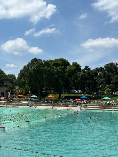 This image shows a large, outdoor swimming pool in Naperville, Illinois, USA. The pool is filled with clear, turquoise water and has lane ropes marking swimming lanes. Several people are visible swimming or wading in the pool. Around the pool, there are grassy areas with people sunbathing and relaxing under colorful umbrellas. A stone building with a brown roof is visible on the left side of the image, and lush green trees form a backdrop. The sky is blue with scattered white clouds.