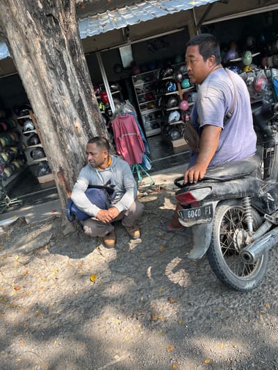 A man is sitting on a motorcycle, looking away from the camera. Another man is squatting next to a tree, with a backpack at his feet. The scene appears to be in front of a shop selling helmets, with shelves of helmets visible in the background. The ground is rough, a mix of gravel and stone, with scattered leaves. The overall environment suggests a street-side setting in a warm, possibly tropical climate, consistent with Kludan, Indonesia. The lighting indicates it is daytime, with dappled sunlight and shadows.
