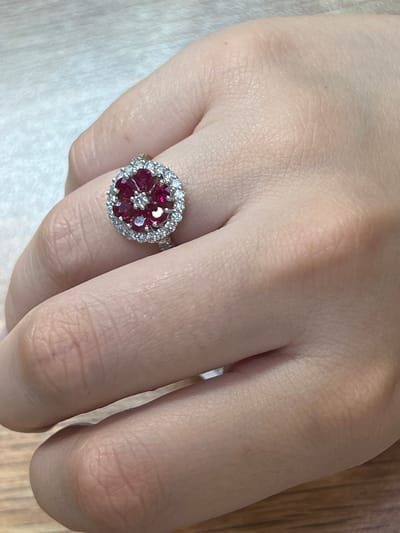 A close-up, high-angle shot shows a person's left hand adorned with a ring. The ring features a central cluster of deep red, marquise-shaped gemstones, possibly rubies, arranged in a floral pattern. This central design is encircled by smaller, clear, round-cut gemstones, likely diamonds, which form a halo. The ring is set on a silver or white gold band. The person's skin is fair and shows the natural texture of fingers and knuckles, with some subtle lines and creases. The background is softly blurred, with a light-colored textured surface on the left and a warm, wood-grained texture on the right. The lighting appears to be soft and natural, suggesting an indoor setting with ambient light. No text is visible in the image. The focus is entirely on the hand and the detailed craftsmanship of the ring.
