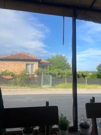 The image depicts an outdoor rural scene in Konstantinovo, Bulgaria, viewed from a sheltered interior. The top of the frame shows a beige fabric canopy structure with visible attachments; a dark brown strap with metal eyelets hangs vertically from the canopy on the upper left. A dark brown vertical post runs down the right side of the image, dividing the view. The immediate foreground, at the bottom, displays the top of a dark wooden or metal bench-like structure. Below this, three potted plants are visible: one with vibrant green foliage, and two with less distinct, possibly dried, vegetation, resting on a gravel surface.
Beyond the viewing pane, an asphalt road traverses horizontally across the middle ground. A narrow strip of green grass borders the road, followed by a dark mesh fence that extends across the scene. To the left, a single-story house with red brick walls and a red tiled hip roof is visible. The house features a prominent white-framed window with horizontal blinds. Green vines and bushes partially obscure the lower portion of the house and grow along the fence line. A small, dark green metal gate or utility box is positioned against the fence to the right of the house. Large, dense green trees populate the background behind the fence and house. The upper portion of the scene reveals a bright blue sky with scattered white clouds. Faint hills or distant terrain are visible on the horizon to the far right. No individuals or active subjects are present.
