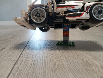 The media file shows a close-up, low-angle shot of a LEGO Technic racing car model positioned upside down on a light-colored wooden floor. A LEGO minifigure, dressed in blue overalls with red and yellow accents on its legs, stands directly beneath the car, seemingly supporting it from below. The car is white with red and black racing stripes and features the text "RACING CAR" and "GTR" on its side. Silver LEGO wheels are visible. The background is out of focus, showing a light-colored wall and a doorframe, suggesting an indoor setting, likely a home. The lighting appears to be artificial and is casting shadows on the floor, indicating it is likely daytime but the specific time is not clear. The scene evokes a playful and imaginative scenario, perhaps a child playing with their LEGO set. The location context is Oleksandriia, Ukraine.