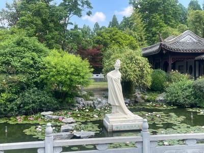 The image depicts a serene Chinese garden in Hailing District, China. A white statue stands prominently in the center of a pond filled with water lilies and scattered rocks. The pond is surrounded by lush greenery, including trees and bushes, with a traditional Chinese pavilion visible in the background to the right. A stone bridge is partially visible in the distance. In the foreground, a stone railing adds to the scene's classical aesthetic.