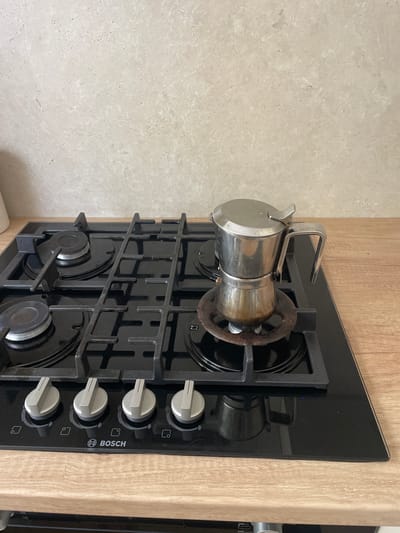 A stovetop coffee maker is situated on a black glass gas stove with a chrome finish and wooden countertop. The coffee maker is made of stainless steel and is on the right side of the stovetop. The stovetop has four gas burners and four control knobs with indicator symbols. The background is a light-colored tiled wall. The scene is set indoors, likely in a kitchen. There is no indication of the city of Cahul, Moldova in the image.