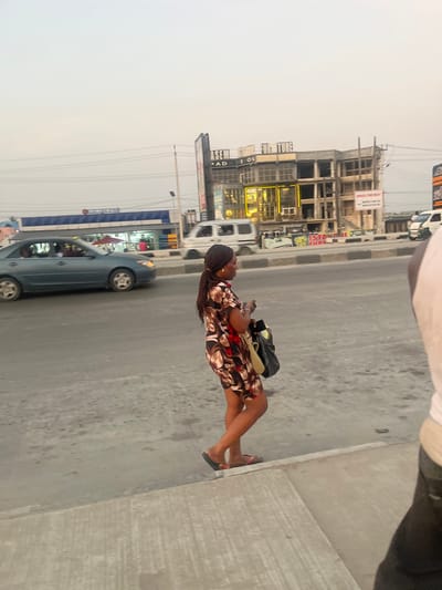 An outdoor shot depicts a woman in a floral dress standing on a sidewalk, facing a street with passing cars. A large building with glass windows stands nearby, and a gas station is visible in the background. The sky is overcast, and the overall scene suggests an urban setting with a mix of commercial and residential elements.