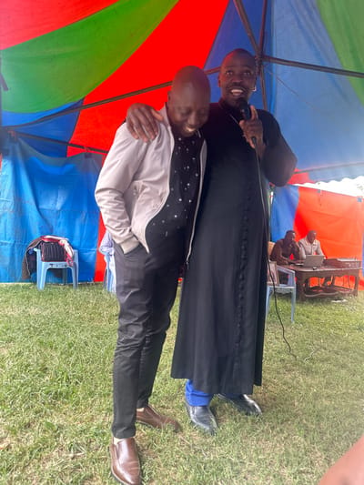 The image captures an outdoor event in Kiganjo, Kenya, under a vibrant, multi-colored tent. In the foreground, two men are prominently featured. One man, on the left, wears a long, dark, buttoned robe, possibly clerical attire, and holds a microphone close to his mouth. He is smiling broadly, conveying cheerfulness or engagement with an audience. His shoes are black, and blue fabric is visible peeking from under his robe.

The second man, positioned slightly in front and to the right, is embracing the robed man. He is wearing a light-colored jacket over a dark shirt with subtle white patterns, and dark pants, with brown shoes. He also appears to be smiling or laughing, showing warmth and camaraderie.

In the background, beneath another section of the tent, other individuals are visible with what appears to be sound or musical equipment, including a keyboard or synthesizer, suggesting a performance or presentation is taking place. The ground is covered in green grass, indicating an open-air setting. The lighting suggests it is daytime. No visible text is discernible in the image.
