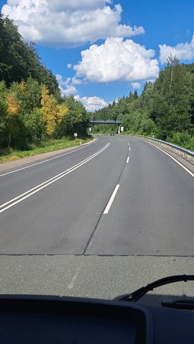 The image shows a view from inside a vehicle driving on a paved road in a forested area. The road curves to the right and has two white lines in the center, indicating a two-lane road. There is a guardrail on the right side of the road. In the distance, there is a bridge crossing over the road, and a few cars are visible on the bridge. The sky is blue with white, fluffy clouds. The trees on either side of the road are green, with some yellow foliage on the left side, suggesting it may be early autumn. The foreground shows the dashboard and windshield wipers of the vehicle. This scene is consistent with a road in the vicinity of Burbach, Germany, a town known for its scenic natural surroundings and forested landscapes.