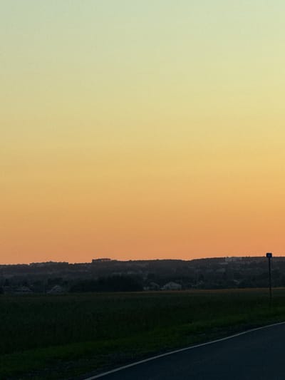 The image depicts a landscape scene at dusk or dawn, with a gradient sky transitioning from pale yellow at the top to a warm orange near the horizon. In the middle ground, a dark, silhouetted line of trees and buildings stretches across the frame, suggesting a rural or suburban area. A dark green field occupies the foreground, with a dark asphalt road curving into the lower right corner. A small blue sign with a white number "4" is visible on a pole to the right of the frame. The location is identified as Morozovka, Russia. There are no people or animals visible in the image.