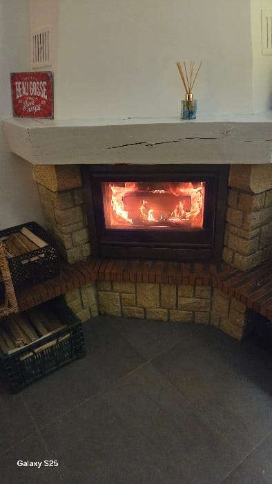 An interior scene, presumed to be within a residence in Ottonville, France, depicts a fireplace as the central subject. The fireplace features a glass-fronted insert containing actively burning orange and yellow flames. The fireplace surround is constructed from light brown, roughly cut stone blocks, with a lower section and hearth trimmed in reddish-brown bricks. A thick, distressed white wooden mantelpiece rests above the fireplace opening.
On the left side of the mantelpiece, a rectangular red sign with white text, partially legible as "BEAU GOSSE," is angled against the white wall. To the right of the sign, a small, patterned blue glass bottle with an orange metallic neck holds several natural reed diffuser sticks. Above the sign, a white rectangular ventilation grille is visible on the wall.
To the left of the fireplace, two dark-colored containers filled with split firewood are stacked. The bottom container is a green plastic crate, and the top is a dark-colored woven basket. The floor in the foreground consists of large, dark gray square tiles with visible grout lines. No individuals are present in the image. The environment is an enclosed domestic space.