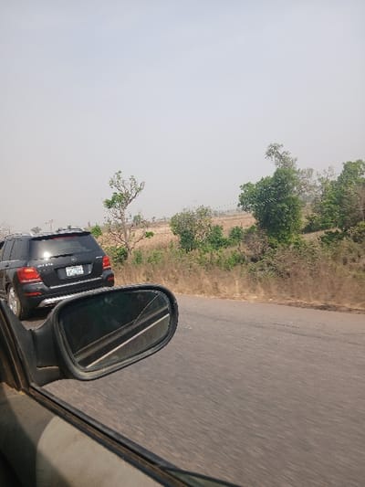 The image depicts a daytime scene on an asphalt road in Bokana, Nigeria, as observed from a moving vehicle. In the immediate foreground, the exterior left side mirror of the capturing vehicle is visible, featuring a crack across its upper left glass surface. Ahead on the road, a dark-colored Mercedes-Benz GLK 350 SUV is traveling in the same direction. The SUV's rear is visible, displaying a Nigerian license plate inscribed with "LAGOS GGE-305 HH". The road is dark grey and appears to be a two-lane thoroughfare. To the right of the road, the landscape consists of dry, light brown vegetation, scattered trees with varying degrees of foliage, and distant fields under a hazy, light grey sky. No individuals are visible.