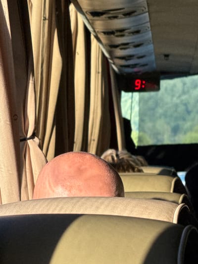 This media file shows a close-up view of a person's head and shoulder, positioned behind upholstered seating. The scene appears to be indoors, possibly within a vehicle such as a van or RV, given the presence of the seating and a visible power strip with outlets. A digital clock on the power strip displays "9:6", indicating the time. Sunlight is filtering in from what seems to be a window or windshield, creating strong shadows across the striped fabric of the seating. The overall lighting suggests it is daytime. The image is taken from a low angle, emphasizing the texture of the fabric and the curve of the person's head. A hint of a cat's head is visible in the background, peeking out from behind the seating, adding a domestic element to the scene. The focus is on the textures and the interplay of light and shadow, with the person's skin appearing smooth and slightly flushed in the sunlight.