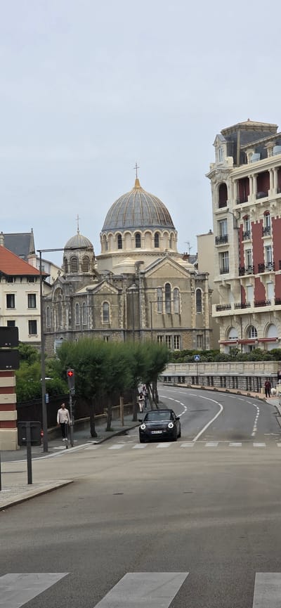 This image captures a street scene in Biarritz, France, dominated by the Saint Alexander Nevsky Russian Orthodox Church.
In the mid-ground and background, the prominent Saint Alexander Nevsky Russian Orthodox Church stands with its light-colored stone facade. It features a large central dome and a smaller secondary dome, both ribbed and topped with golden spires bearing Orthodox crosses. The church's architecture includes multiple arched windows and decorative elements. To the immediate right of the church is a grand, multi-story building with a light-colored facade, ornate balconies, classical columns, and sections painted in a deep red. Its roof is dark and mansard-style. To the left of the church, a smaller residential building with a reddish-brown tiled roof and white walls is visible.
The foreground features a paved, asphalt road that curves to the right, leading uphill. A white, striped pedestrian crosswalk is marked on the asphalt in the bottom left. A black convertible Mini Cooper with its headlights illuminated is positioned on the road, facing the viewer, slightly right of the center. Its license plate, "GR 271-TE," is visible.
Along the left side of the road, a sidewalk is present, bordered by a dark fence and a row of pruned, green trees. A traffic light showing a red signal is mounted on a pole near the sidewalk. A man wearing a light-colored shirt and dark trousers stands on the sidewalk near this pole. Further down the street on the sidewalk, several other pedestrians are visible, one appearing to walk a small white dog. A white pedestrian crossing sign (blue circle with white figure) is visible on the right side of the road further up the curve. The sky is overcast with a uniform light gray tone.