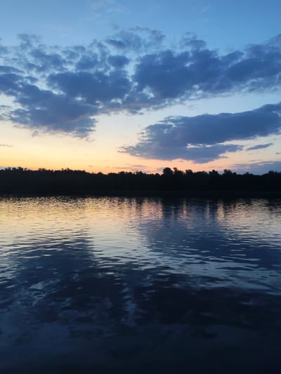 The image captures a twilight scene over a body of water, likely a lake or a wide river. The sky is a gradient of blue at the top, transitioning to orange and yellow near the horizon. Clouds are scattered across the sky, with darker, bluish-gray formations dominating the upper portion.
A dark, silhouetted treeline stretches across the horizon, separating the sky from the water. The water surface is rippled, reflecting the colors of the sky and the trees, creating a slightly blurred and mirrored effect.
There are no visible people or other identifiable objects in the scene. The setting appears to be a natural environment, possibly on the outskirts of Ermolino, Russia, though specific landmarks are not present to confirm this definitively. The focus is on the natural landscape and the interplay of light and reflection.
