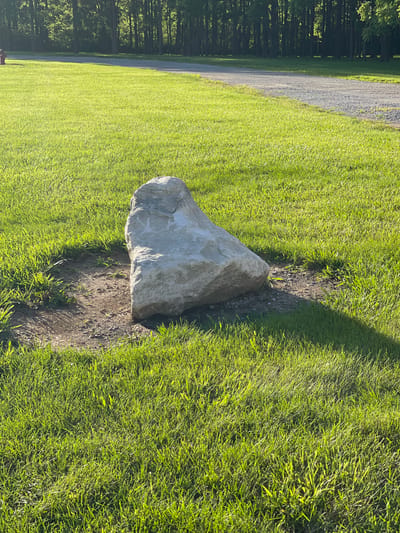 A large, grey, triangular rock sits in a patch of bare earth surrounded by bright green grass. The rock is lit by sunlight, casting a shadow to the right. In the background, a gravel path curves to the left, bordered by more green grass and a dense line of dark trees. The scene is outdoors, likely in a park or natural area. There are no people or animals visible in the image. The location is identified as Eaton, United States of America.