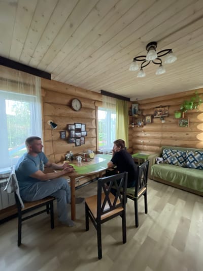 In a room with wood-paneled walls and ceiling, a man and a woman are seated at a wooden table. The man, wearing a blue t-shirt and gray pants, is on the left, facing right. The woman, in a black shirt, is on the right, facing left. A green placemat is on the table between them, along with various items including bottles and a small white cup. Two wooden chairs with black frames are positioned at the table. The room contains a sofa with patterned cushions on the right, a wall clock and a cluster of framed photos on the back wall, a plant on a shelf, and windows with sheer curtains on the left. The flooring is light-colored wood planks. This scene appears to be in a residential dwelling located in Kumyonys, Russia.