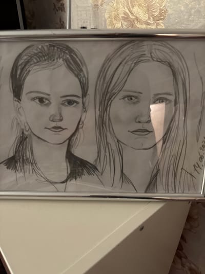 A black and white pencil sketch of two young women is framed in a silver metal frame. The sketch is on paper and is covered by glass. The left woman has her hair pulled back into a ponytail and is wearing earrings. She is looking directly at the viewer. The right woman has long, straight hair and is also looking at the viewer. A bright light is shining across the right side of her face. On the right side of the sketch, a date is written in what appears to be pencil: "14.06.2023". The sketch is positioned on top of a white piece of furniture, possibly a shelf or a dresser. The background behind the framed sketch is out of focus, but appears to be a wall with floral wallpaper. The overall setting suggests an indoor environment. The sketch is not located in the city of Pohreby, Ukraine; this information is not present in the image.