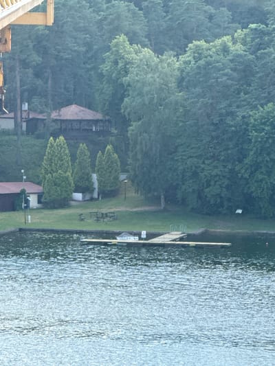 This image depicts a tranquil lakeside scene in Mikołajki, Poland. The primary subjects are the natural elements: a dense, lush forest that forms the backdrop and a calm body of water occupying the right side of the frame. A wooden pier extends into the water, suggesting a recreational area.
On the grassy bank between the forest and the water, there are several man-made structures and objects. A building, possibly a cabin or boathouse, is partially visible amidst the trees. Further along the shore, a few picnic tables and benches are arranged, hinting at a place for relaxation or gatherings. A white square sign is placed on a pole on the grass. The overall setting appears to be a natural park or a lakeside resort.
The lighting suggests it is either early morning or late afternoon, with diffused light and a slightly overcast sky. The water's surface is rippled, indicating a gentle breeze. The atmosphere is serene and peaceful, evoking a sense of calm and natural beauty. There is no visible activity of people in the image, reinforcing the quiet nature of the scene. The time of day appears to be daytime.