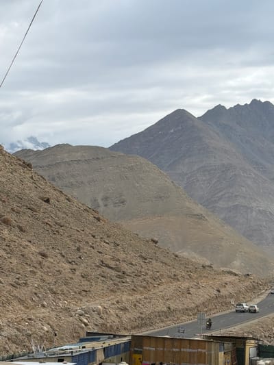 In Chumkar, India, a mountainous landscape features a road with vehicles. A string stretches across the cloudy sky from the upper left corner of the image. To the left, a steep, rocky hill rises, covered in loose stones and dirt. Further back, a wider mountain range with layered rock formations dominates the background. A paved road curves through the scene, with two white cars and a motorcycle visible. In the foreground, a low-rise structure with a corrugated metal roof and various walls is partially visible on the right. The sky is overcast with diffused light.
