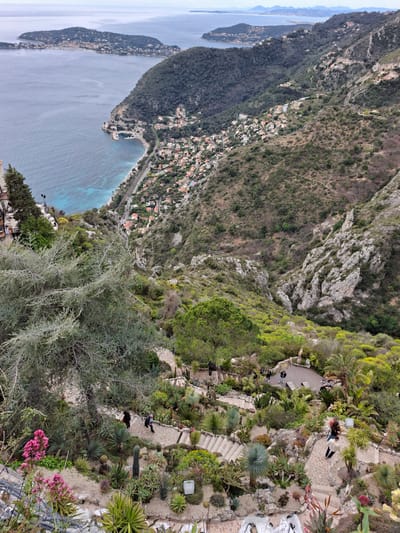 The image captures a high-angle view in Èze, France, overlooking the Mediterranean Sea. The foreground features a garden on a cliff, with pathways and steps winding through cacti and other vegetation. Several people are visible on the paths. Below the garden, the coastline is lined with buildings. On the left, a peninsula extends into the sea. The sky is overcast.