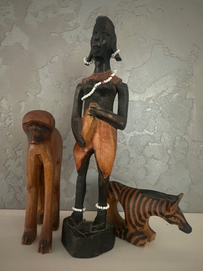 In a domestic setting, three carved wooden figurines are displayed on a white shelf against a textured grey wall. On the left is a brown wooden figurine of a monkey in a standing position. To its right is a taller, dark-skinned figurine of a woman adorned with white and red bead necklaces and white bead bracelets around her ankles and ears. The woman is wearing a short, orange-brown skirt. To the right of the woman is a carved wooden zebra figurine with black stripes on a brown body. The overall scene depicts decorative items arranged for display.