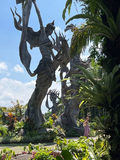 The image shows statues at the "Tirta Empul Temple" in Kedewatan, Ubud, Gianyar Regency, Bali, Indonesia. The gray, stone statues are adorned with traditional Balinese decorations and are surrounded by lush tropical vegetation. A woman in a pink dress stands near one of the statues. The sky is blue with scattered clouds.