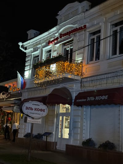 The image depicts a nighttime view of a building facade in Kislovodsk, Russia. The building features ornate architectural details, including pilasters and decorative molding. A balcony on the second floor is adorned with numerous warm-toned string lights. A Russian flag is visible on the left side of the balcony.
On the ground level, there are storefronts with awnings. One awning, on the right, has "ЭЛЬ КОФЕ" (El' Kofe) written on it in white letters. Below this, a sign in a distinctive white oval shape also reads "КОФЕЙНЯ ЭЛЬ КОФЕ КОНДИТЕРСКАЯ" (Coffeehouse El' Kofe Confectionery). Another sign below this indicates "Курортный бульвар №6" (Resort Boulevard No. 6).
To the left, a storefront with the partially visible sign "банда" (banda) is present. Two individuals are visible near this entrance, one wearing a dark suit and the other a white shirt and dark trousers.
The scene is illuminated by artificial lighting, casting a warm glow on the building and its decorations. The overall atmosphere suggests a commercial area in the evening.