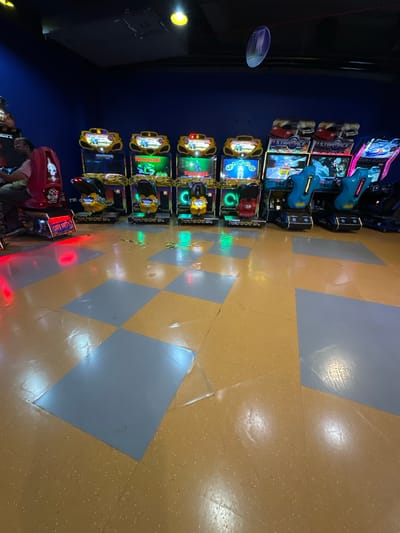 The image depicts a row of arcade games in an indoor setting, likely an entertainment center in Kalyan-Dombivli, India. The arcade games are arranged along a wall with blue paint. There are several games that appear to be motorcycle racing games, followed by a pair of blue gun-themed racing games near the end of the row. The floor has a diamond pattern with yellow and grey tiles. People are visible near the bottom of the frame.