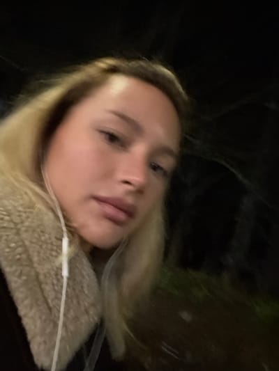 A close-up, dimly lit, slightly blurred selfie depicts an individual, identified as female, with blonde hair and a fair complexion, looking towards the camera. White wired earphones are visible, with the wire extending downwards from her left ear. She is wearing a light-colored, likely beige or cream, fur-lined or sherpa-style jacket or coat collar. The background is predominantly dark and out of focus, revealing subtle hints of green foliage or ground cover in the lower right, indicative of an outdoor setting during nighttime or in a poorly lit environment. The scene is located in Fontenay-sous-Bois, France.
