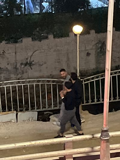 A photograph taken outdoors in the evening in Dummar Municipality, Syria, shows two people embracing near a set of metal stairs and a lamppost. The person on the left appears to be wearing a dark-colored jacket and light-colored pants, and has short dark hair. The person on the right appears to be wearing dark-colored clothing. The staircase is silver or light-colored metal and is located near the center of the photo. The lamppost is on the left, with a glowing light bulb. Vegetation is visible on the left side of the photo, and part of a structure or building is visible in the background behind the couple. A metal bar is visible in the foreground at the top and right of the image.