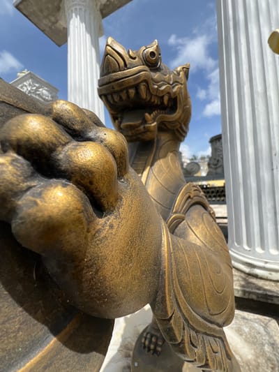 A close-up, low-angle shot features a golden, stylized dragon statue in the foreground. The statue has a detailed head with sharp teeth and prominent eyes, and its front paw, with visible toes, is positioned prominently on the left side of the frame. The body of the dragon is partially visible, showing intricate scale-like patterns and ornate shoulder armor.
In the background, two large, fluted white columns dominate the scene, suggesting a classical or temple-like architectural setting. The sky above is a bright blue with scattered white clouds. Further in the background, other architectural elements, possibly more statues or structures, are visible, hinting at a larger complex or garden.
The image was captured in Pa Khlok, Thailand, as indicated by the contextual information. The lighting suggests it is daytime, with the sun casting shadows and highlighting the metallic sheen of the dragon statue. The overall impression is one of ornate, possibly spiritual or decorative artistry within an open-air environment.