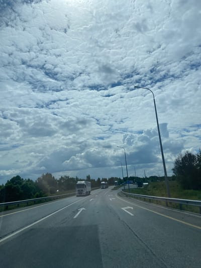 A view from a vehicle traveling on a highway with multiple lanes. The sky is filled with dramatic, white, puffy clouds against a light blue background. Streetlights with curved poles line the side of the road. On the right side of the road, there is a metal guardrail and grassy embankment with trees and foliage. In the distance, several trucks are visible on the highway. The road has white lane markings and directional arrows painted on its surface. The scene suggests a journey on a cloudy day in a semi-rural or suburban area. The location is identified as the city of Pervomaiskoye, Russia, implying it's a road within or near this city.