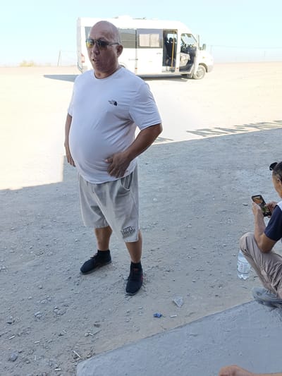 A man stands in a dusty outdoor area near a white van. He is wearing a white t-shirt, grey shorts, black socks, and black sneakers. He has on sunglasses and is holding his right hand on his stomach. The ground is covered in sand and small pebbles. In the background, there is a white van with its door open and windows covered. Beyond the van, the landscape appears arid with some sparse vegetation and utility poles under a clear sky. To the right of the frame, another person is partially visible, crouching and looking at a smartphone. A plastic water bottle is on the ground near this person. The scene suggests a location in a dry, possibly rural or industrial area, consistent with parts of Dosan, Kazakhstan.