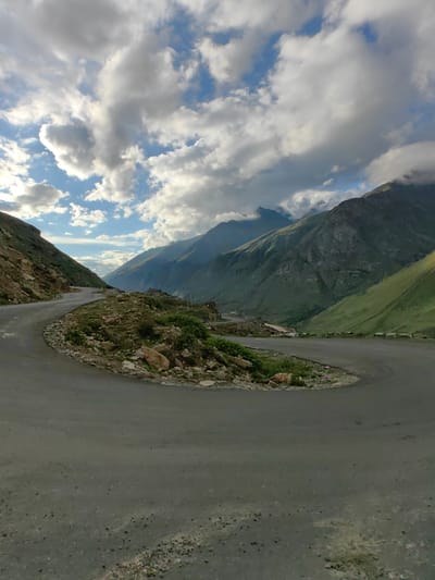 The image depicts a winding asphalt road in a mountainous region, likely Gramphu, India, under a partly cloudy sky. The road curves sharply in the foreground, with rocky terrain and sparse vegetation on its edges. In the background, majestic green and rocky mountains rise, with some peaks shrouded in clouds. The lighting suggests it might be late afternoon or early morning. There are no people or vehicles visible in the immediate foreground, but a small white vehicle can be spotted in the distance on the road, further down the valley. The overall scene is one of a remote, rugged, and scenic mountain landscape.