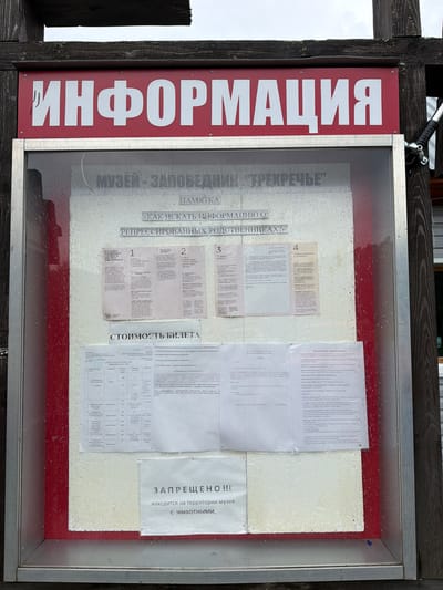 The image shows an information board at the "TREKRECHYE" Museum-Reserve in Ust'-Kabyrza, Russia. The board is mounted on a wooden post and has a red frame. The top of the board displays the word "ИНФОРМАЦИЯ" (INFORMATION) in large white letters. Below this, there is text in Russian indicating that it is a memorial and instructions on how to find information about repressed relatives. There are four sections numbered 1 to 4, each detailing a step in the process. Further down, a section titled "СТОИМОСТЬ БИЛЕТА" (TICKET PRICE) lists different ticket categories and their prices. At the bottom of the board, a prominent "ЗАПРЕЩЕНО!!!" (FORBIDDEN!!!) message in large capital letters is followed by text stating that animals are not allowed on the museum territory. The board is behind a glass panel which shows streaks of water or condensation. There are no people visible in the image. The environment appears to be outdoors, with wooden structures and a cloudy sky in the background.