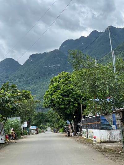 The image depicts a street scene in Lùng Tám Commune, Vietnam, characterized by a paved road leading towards lush green mountains under a cloudy sky. On the left side of the road, there are several motorcycles parked, with a few individuals standing or sitting near them. A young child in a red shirt is standing near the motorcycles. Further down the road, more motorcycles and people are visible, suggesting a tranquil, rural atmosphere. The right side of the road features a fence and some structures, with a signpost marked "H1 58" visible. Large trees with dense foliage line both sides of the road, and the mountainous backdrop is covered in vegetation. The overall impression is of a quiet, developing area integrated with the natural landscape.